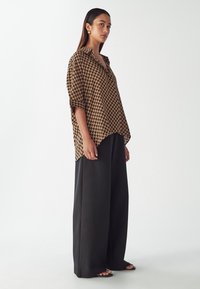 Brązowa koszula w wzory z czarnymi geometrycznymi kształtami, z kołnierzykiem i podwiniętymi rękawami, w zestawieniu z czarnymi spodniami o szerokich nogawkach. Gładka tekstura tkaniny.