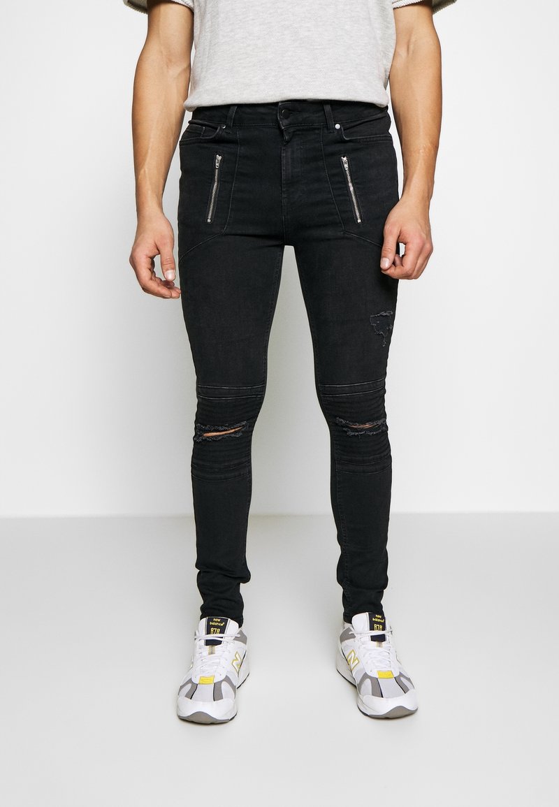 Jeans skinnies negros con bolsillos con cremallera, que presentan detalles desgastados y un corte ajustado. El tejido tiene una textura suave con costuras visibles.