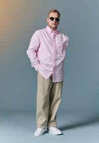 Light pink striped button-up shirt, beige pants, and white sneakers. Round sunglasses complete the casual outfit. Smooth fabric texture.