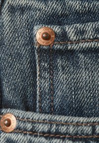 Ύφασμα τζιν σε διάφορες αποχρώσεις του μπλε, με χάλκινα καρφιά και ορατή ραφή, τονισμένο από λεπτομέρεια τσέπης.