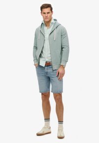 Lichtgroene zip-up hoodie met trekkoorden, gestreepte shirt, blauwe denim shorts, roomkleurige sneakers en beige sokken met blauwe strepen.
