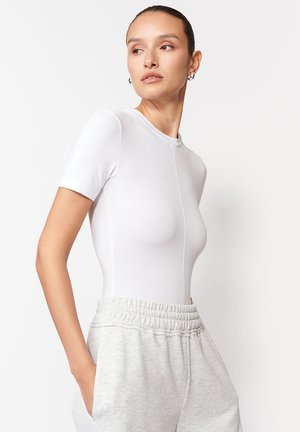 Fehér, rövid ujjú bodysuit, testhez simuló kialakítással, varrás nélküli textúrával és kerek nyakkivágással, szürke, húzózsinóros sportnadrággal párosítva.