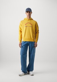 Žltá bavlnená mikina s červeným textom "HILFIGER EST. 1985". Skombinované s modrými širokými cargo džínsami a šedými teniskami. Nastaviteľná čiapka dotvára vzhľad.