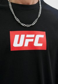 Sort bomulds T-shirt med et stort rødt rektangulært grafisk design med hvid "UFC" tekst. Inkluderer et sølvfarvet kædede armbånd.