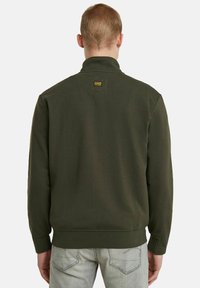 Felpa verde oliva, tessuto di cotone testurizzato, colletto alto, polsini e fondo a costine, patch di marca visibile sul retro.
