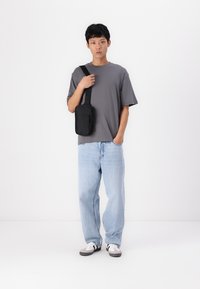 Tricou gri cu mânecă scurtă, blugi bleu deschis, cu o croială lejeră, și geantă de umăr neagră. Sneakers casual cu accente albe și gri. Fundal simplu.