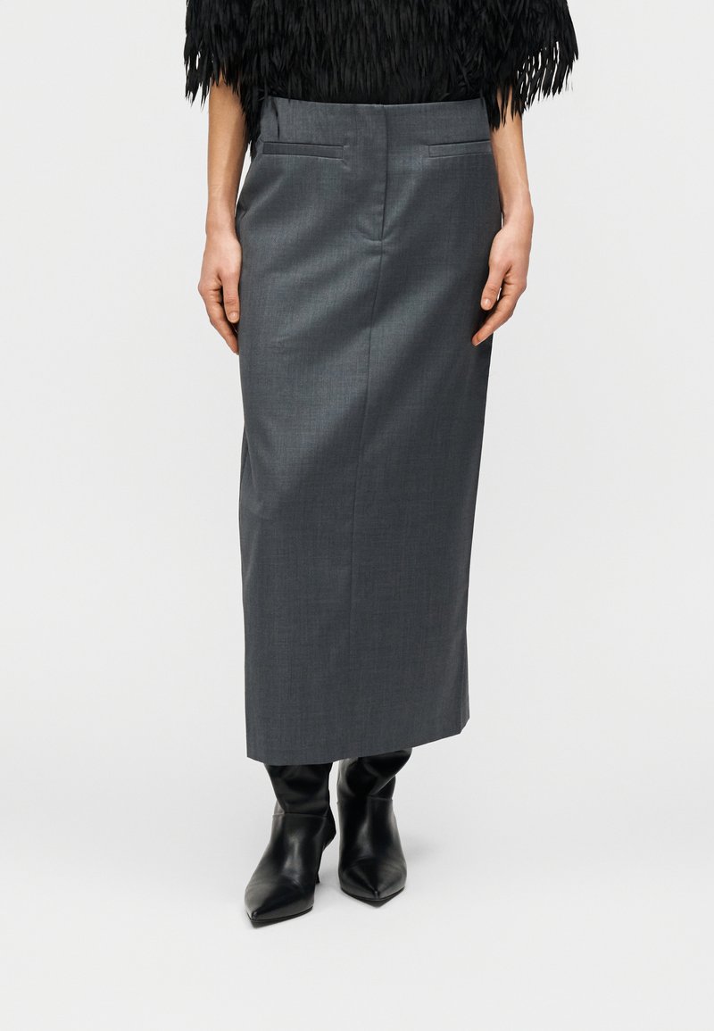 Jupe crayon grise sur mesure avec ceinture, poches latérales et texture lisse, assortie à des bottines noires à bout pointu.