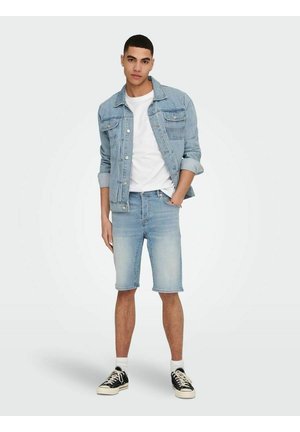Lys blå denimjakke, hvid skjorte, lys blå denimshorts, sorte sneakers med hvide såler, sat mod en ensfarvet baggrund.