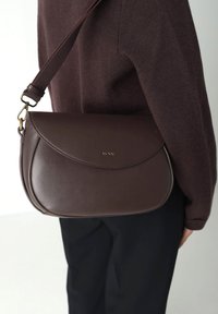 Persona che indossa un maglione marrone scuro e pantaloni neri, mentre porta una borsa a tracolla in pelle marrone scuro con pattina e hardware dorato.