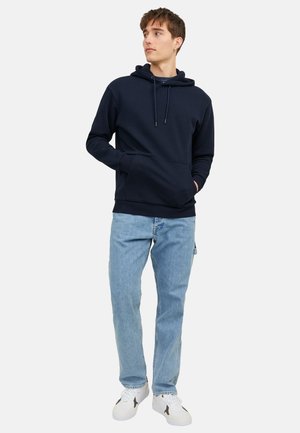 Junger Mann trägt einen schwarzen Hoodie, hellblaue Jeans und weiße Sneaker, steht mit den Händen in den Taschen und schaut zur Seite.