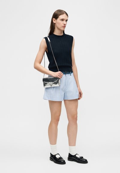 Γυναίκα με μαύρο αμάνικο πουλόβερ, ανοιχτό μπλε ριγέ σορτς, λευκές κάλτσες, μαύρα παπούτσια και κρατάει ασημένια τσάντα ώμου.