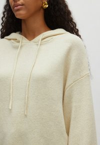 Beige Kapuzenpullover in lässiger Passform, aus weichem Strickmaterial, mit schlichter Gestaltung und Kordelzug im Halsbereich.