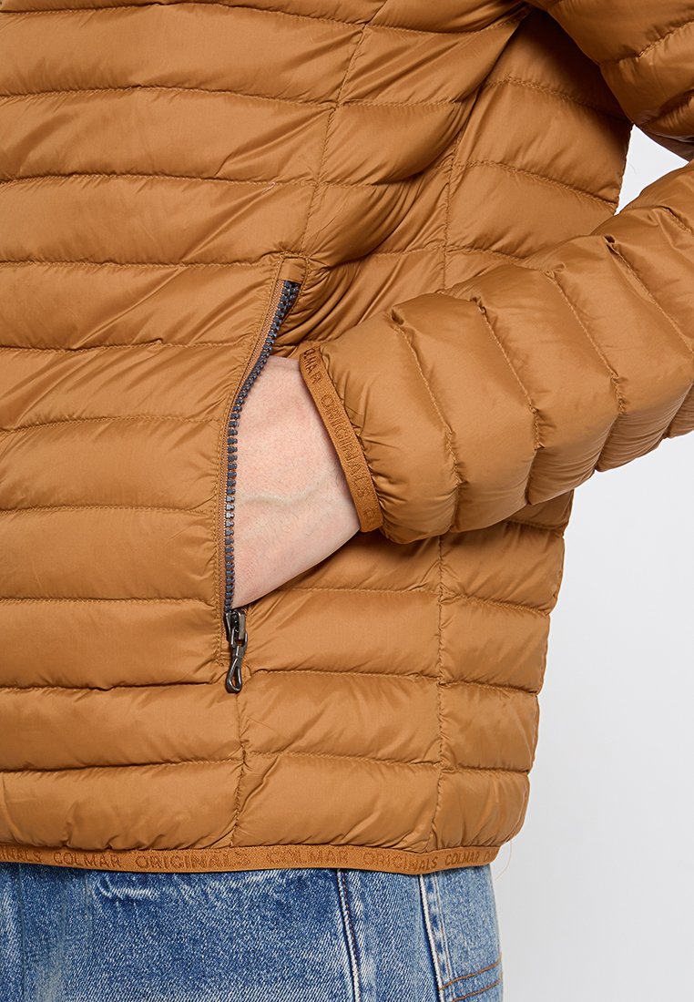 Giacca a vento marrone con motivo a righe orizzontali, tasca laterale con zip e polsini a costine, indossata sopra jeans chiari.