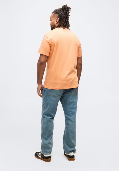 Άνδρας με dreadlocks στέκεται με την πλάτη, φορώντας ροζ μπλουζάκι, μπλε τζιν και μαύρα και λευκά αθλητικά παπούτσια σε απλό λευκό φόντο.