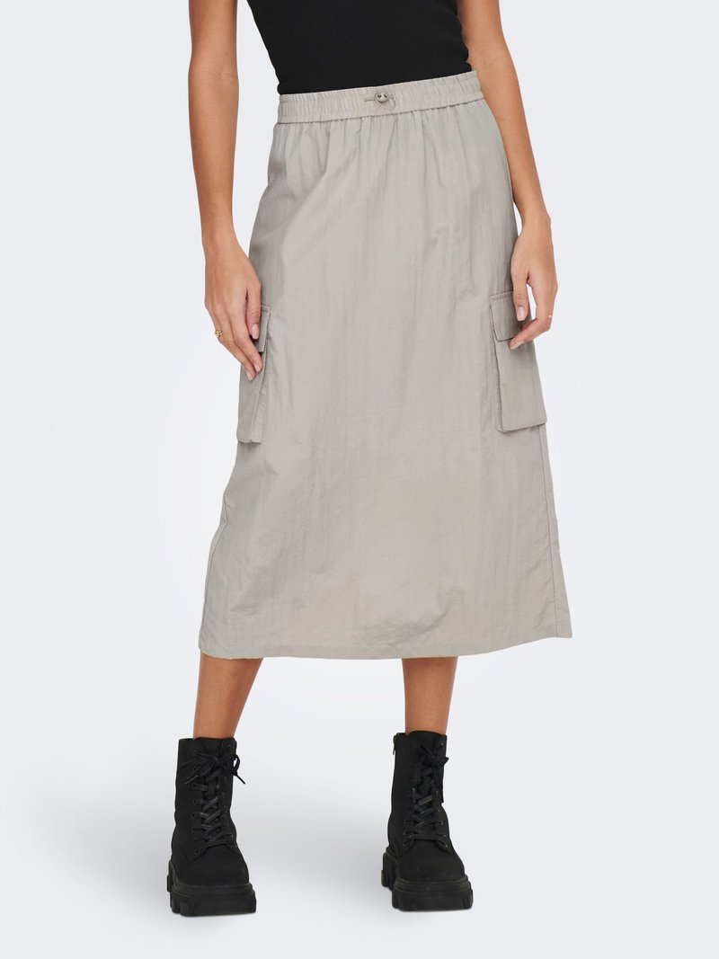 Kvinde iført beige midi cargo-nederdel med elastisk taljebånd og sorte militærstøvler, stående med hænderne hvilende på nederdelslommerne.