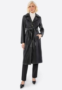 Černý kožený trenčkot s širokými klopami, zapínáním na knoflíky, páskem v pase a dlouhými rukávy. Nosit se přes černé přiléhavé kalhoty a špičaté boty.