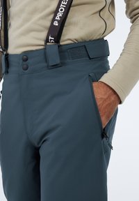 Skibroek in donkerteal stof met een gladde textuur. Voorzien van verstelbare bretels, knoopsluiting, een ritszak en een klittenbandstrap bij de taille.