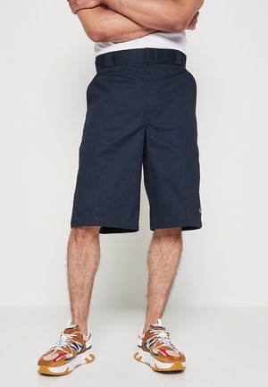 Mann trägt marineblaue knielange Shorts und mehrfarbige Turnschuhe, steht mit verschränkten Armen vor einem schlichten Hintergrund.