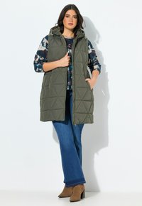 Žena oblečená v olivově zelené prošívané bezrukávové bundě, vzorovaném dlouhém rukávu, modrých džínách a hnědých botách na podpatku, stojí s jednou rukou v kapse.