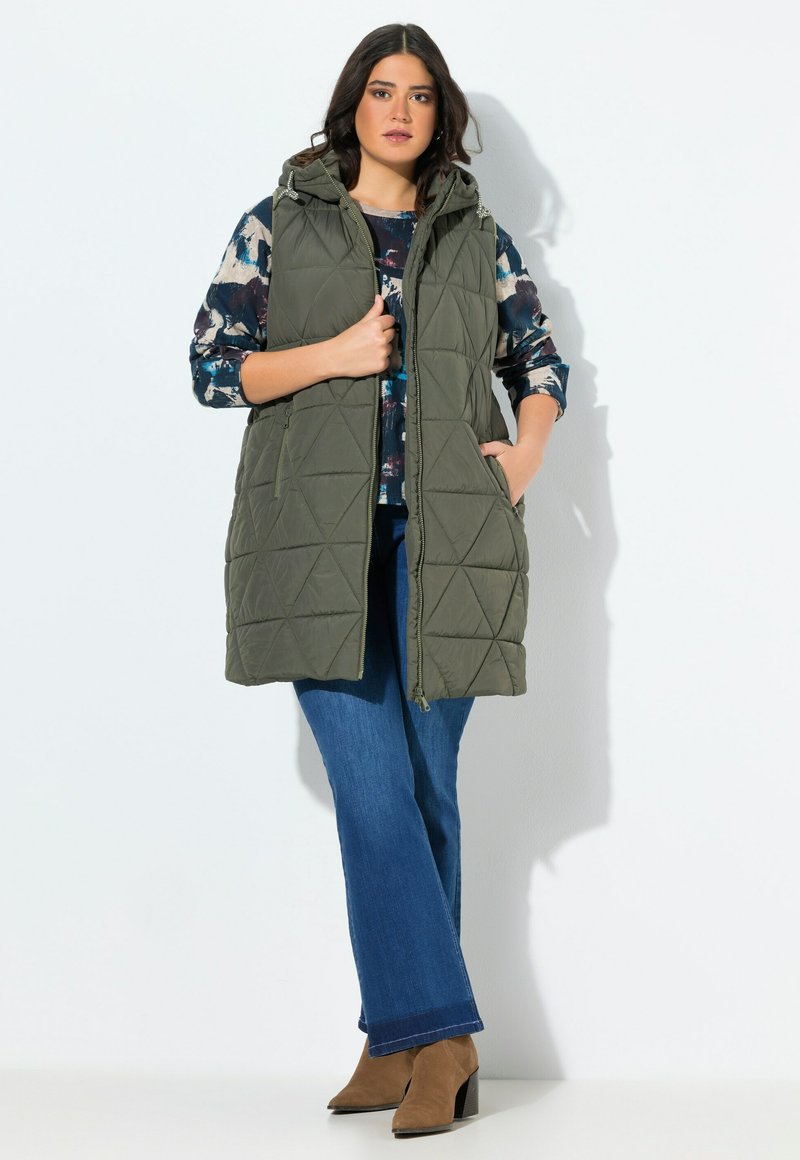 Žena oblečená v olivově zelené prošívané bezrukávové bundě, vzorovaném dlouhém rukávu, modrých džínách a hnědých botách na podpatku, stojí s jednou rukou v kapse.