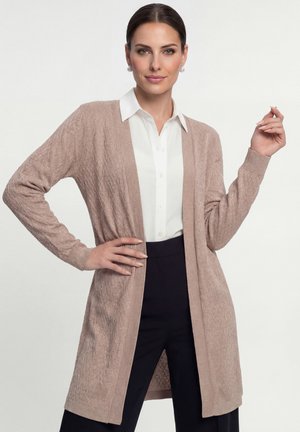 Frau trägt eine hellbraune, strukturierte Strickjacke über einem weißen Hemd und schwarzen Hosen, posiert mit einer Hand an der Hüfte und der anderen erhoben.