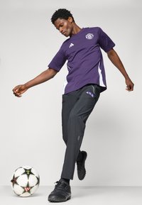 Paarse sporttrui met witte accenten, zwarte sportbroek met een paars logo en zwarte voetbalschoenen, naast een gestreepte voetbal.