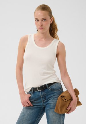 Femme avec queue de cheval blonde portant un haut blanc sans manches, jean bleu avec ceinture noire, tenant une veste marron pliée dans la main droite.