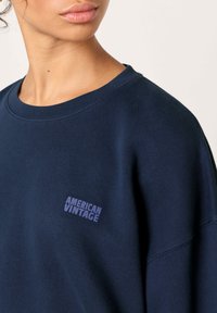 Tamsiai mėlynas džemperis iš medvilnės mišinio, turintis apvalų kaklo iškirptę ir laisvą kirpimą. Ant jo išsiuvinėtas „AMERICAN VINTAGE“ logotipas šviesiai violetine spalva.