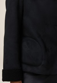 Giubbotto in suede nero con un polsino testurizzato e morbido e una tasca laterale discreta. Il design presenta una finitura liscia e uno stile minimalista.