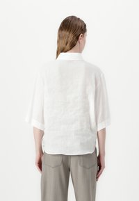 White linen shirt with short sleeves, a small collar, and a relaxed fit. Paired with light gray trousers featuring subtle stitching details.