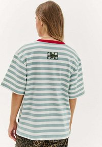 Osoba s dlouhými vlasy, oblečená v modro-bílém pruhovaném tričku s červeným límečkem a květinovou nášivkou na horní části zad, doplněná leopardími kalhotami.