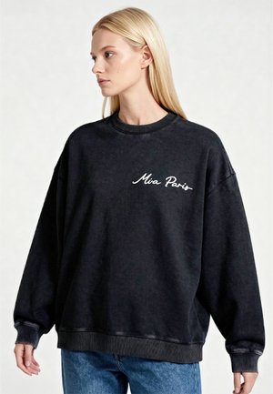 Jonge vrouw met steil blond haar die een zwarte oversized trui draagt met de tekst "Mia Paris" op de borst en een blauwe spijkerbroek.