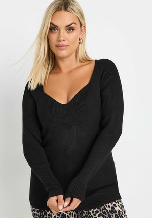 Donna con capelli biondi lisci che indossa un maglione nero a coste con scollatura profonda a V e pantaloni con stampa leopardata, in piedi su uno sfondo neutro.