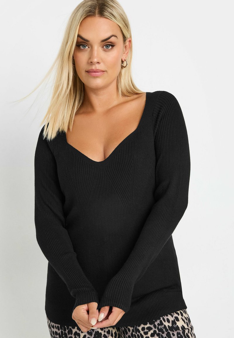 Nainen, jolla on suorat vaaleat hiukset, pukeutuneena mustaan ribbipaitaan, jossa on syvä V-aukko, ja leopardipainatuksellisiin housuihin, seisoo yksinkertaista taustaa vasten.