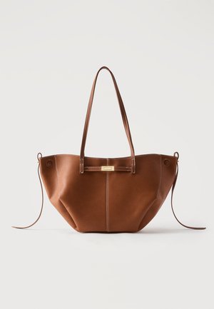 Bruine leren tote bag met lange riemen, decoratieve voorriem met goudkleurige gesp en zijkant met trekkoorddetails tegen een witte achtergrond.