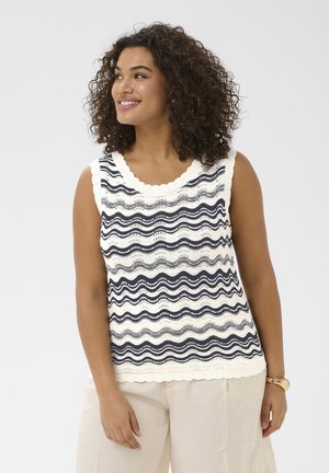 Lächelnde Frau mit lockigem Haar, die ein ärmelloses weißes Stricktop mit marineblauen Wellenstreifen und hellbeige Hose vor einfachem Hintergrund trägt.