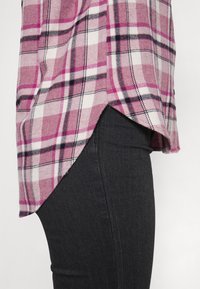 Rutigt skjorta i rosa, vitt och svart; tillverkad av mjukt tyg med långa sidoventiler. Pairad med mörka, tighta jeans.