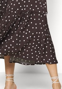 Zwarte midi-rok met witte polka dots, vloeiende stof, gedragen met zichtbare benen en witte wikkelbandjes om de enkels op blote voeten.