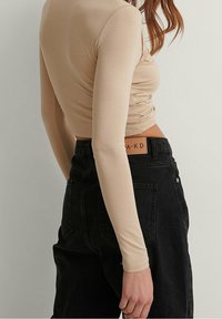 Top beige a maniche lunghe con dettagli arricciati sui lati, abbinato a pantaloni in denim neri a vita alta con etichetta sulla cintura.