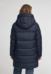 Person som bär en mörk marinblå puffjacka med huva, sedd bakifrån, stående mot en enkel ljus bakgrund.