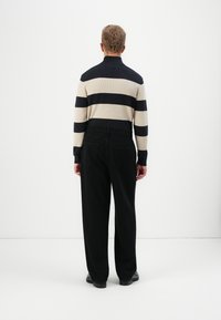 Mann, der einen schwarz-beigen gestreiften Pullover, schwarze Hose und schwarze Schuhe trägt, steht mit den Händen an den Seiten vor einer weißen Wand.