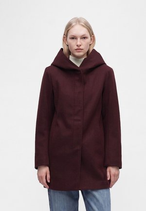 Femme portant un manteau bordeaux foncé avec un col haut et un jean bleu, debout devant un fond gris clair uni.