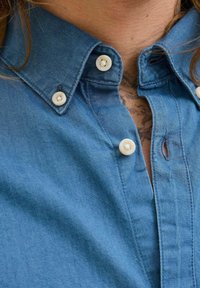 Blå denimskjorta med knapplist, prydd med vita knappar och en liten öppen placket nära nacken. Slät textur och avslappnad design.