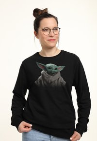 Sort sweatshirt med et grafisk motiv af en grøn, storørret karakter med mørke øjne, iført en grå kappe og hætte. Blødt stof, afslappet pasform.