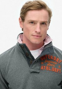 Sudadera gris para hombre con cuello tipo camisa, forro interior rosa claro, detalles de botones y "Superdry" bordado en naranja.
