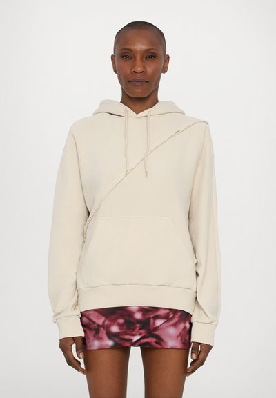 Cremefarvet hættebluse med lomme foran, der har et asymmetrisk design og tekstureret trim detaljer. Sammen med et mønstret lilla nederdel.