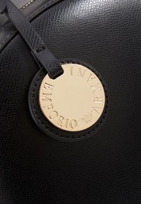 Bolso de cuero negro texturizado con una etiqueta circular de logo dorado con "Emporio Armani" grabado, asegurado por una correa negra.