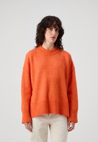 Oransje strikkegenser med ribbet krage og nederkant. Har senkede skuldre og en teksturert overflate. Kombinert med lyse bukser.