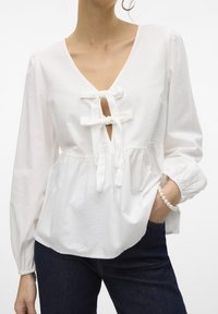 Blusa bianca in cotone con scollatura a V profonda, fiocchi decorativi all'incrocio e maniche lunghe a sbuffo. Presenta un punto vita arruffato e un tessuto strutturato.