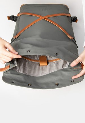 Grijze stoffen rugzak met leren accenten. De open bovenkant onthult een patroon in de binnenvoering en een leren sluiting met riem. Voorzien van drukknoopsluitingen voor extra beveiliging.